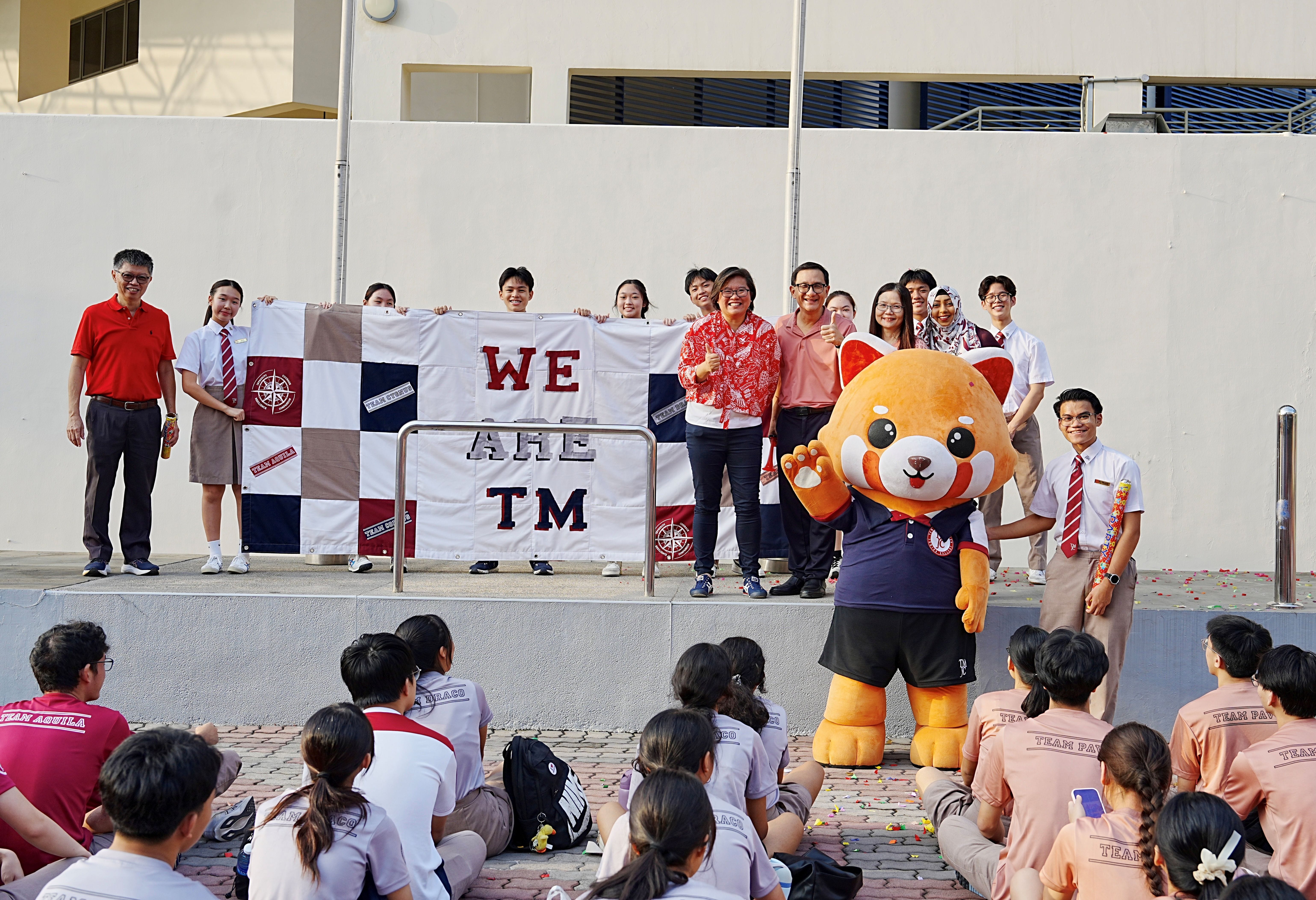 Students and staff proudly display a patchwork banner reading "We Are TM", made from old uniforms, alongside the school's red panda mascot.